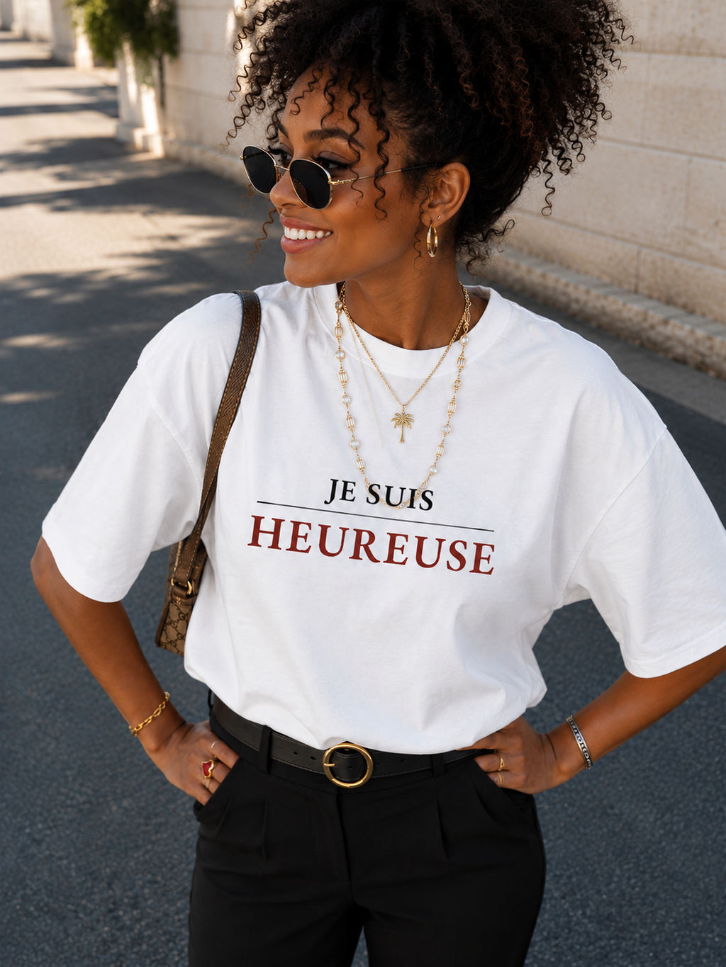 Femme noire souriante portant le t-shirt "Je suis heureuse" de La Femme Entrepreneure avec un pantalon noir et des bijoux dorés dans un look lifestyle en extérieur.